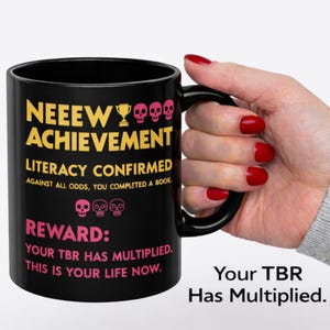 以下が含まれることがあります： 黄色とピンクで「NEEEW ACHIEVEMENT LITERACY CONFIRMED」と書かれた黒いセラミックマグ。マグには「REWARD: YOUR TBR HAS MULTIPLIED. THIS IS YOUR LIFE NOW.」という文字も書かれています。マグは人が持っています。