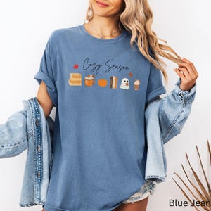 May include: A blue t-shirt with the words "Cozy Season" in script. The shirt features a graphic of fall-themed items, including books, a mug, a pumpkin, a ghost, and a coffee drink. The shirt is a relaxed fit.