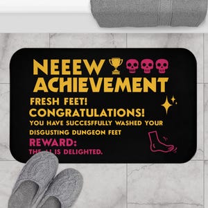 Könnte beinhalten: Eine rechteckige schwarze Badematte mit gelbem Text, der NEEEW ACHIEVEMENT, FRESH FEET! CONGRATULATIONS! YOU HAVE SUCCESFFULLY WASHED YOUR DISGUSTING DUNGEON FEET lautet, und rosa Text REWARD: THE AI IS DELIGHTED, mit Totenkopf- und Pokalsymbolen.