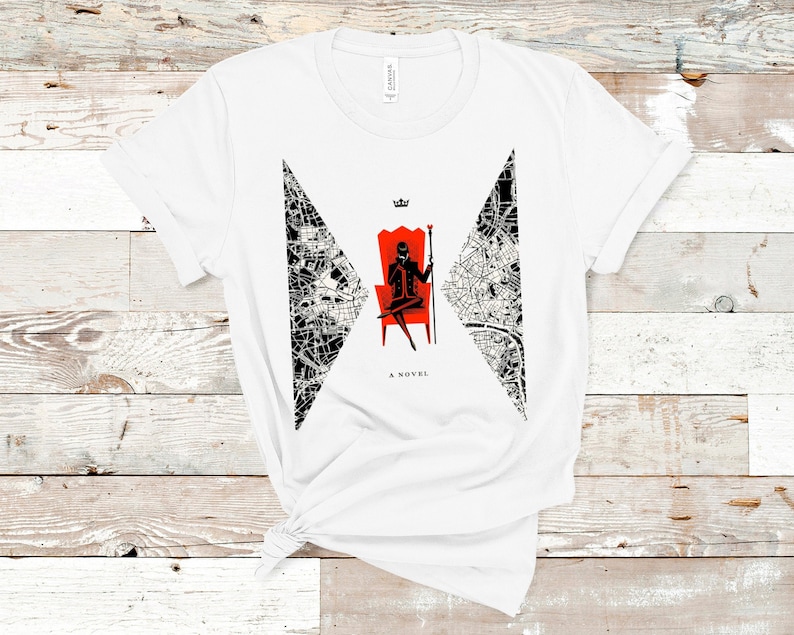 Op de afbeelding: Wit T-shirt met een zwart-witte afbeelding van een stadskaart en een silhouet van een persoon die op een rode troon zit met een kroon erboven. De tekst "A NOVEL" is onder de afbeelding gedrukt.