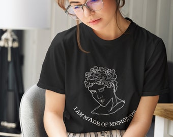 May include: A black short-sleeved t-shirt featuring a white continuous line drawing of a classical bust and the text "I AM MADE OF MEMORIES" printed in a white serif font across the chest area, positioned below the graphic illustration.