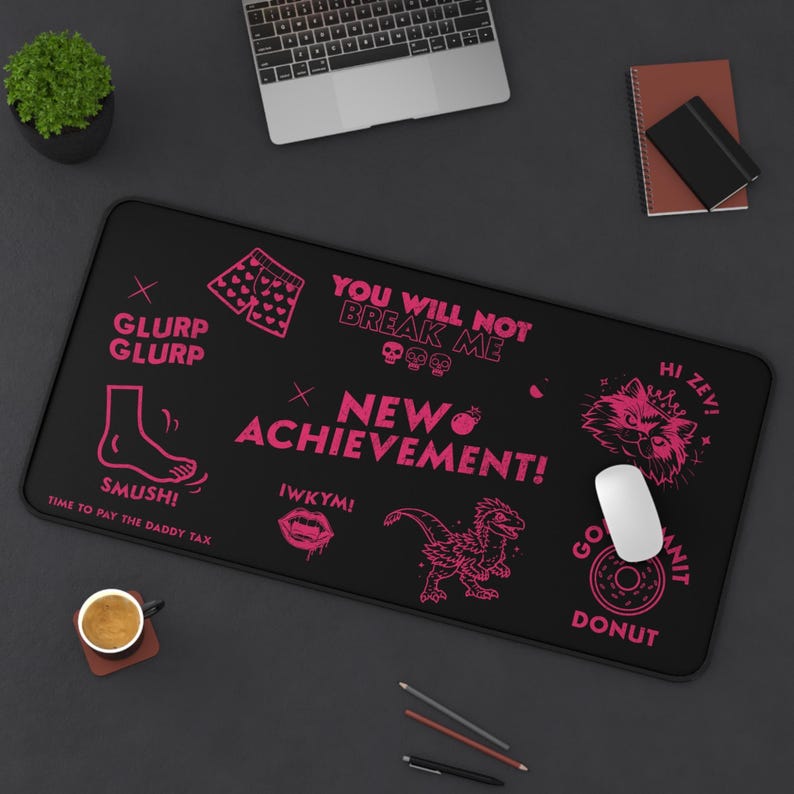 K&ouml;nnte beinhalten: Schwarzes rechteckiges Mauspad mit rosa Grafiken und Text. Das Design umfasst einen Fu&szlig;, einen Dinosaurier, eine Katze und S&auml;tze wie "NEW ACHIEVEMENT!" und "YOU WILL NOT BREAK ME".