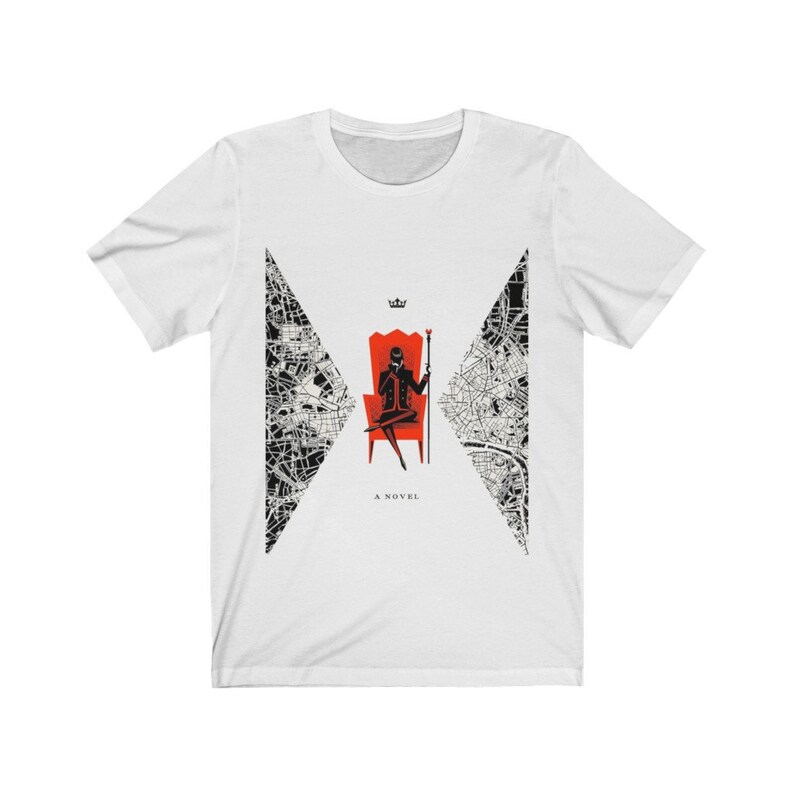 Op de afbeelding: Wit T-shirt met een zwart-wit ontwerp van een stadskaart. Het ontwerp toont een silhouet van een persoon die op een rode troon zit, een staf vasthoudt en een kroon draagt. De tekst "A NOVEL" is onder het ontwerp gedrukt.