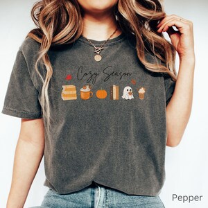 May include: A dark gray t-shirt with the words "Cozy Season" in script. The shirt features fall-themed illustrations including books, a pumpkin spice latte, a pumpkin, a stack of books, a ghost, and a coffee drink. The shirt is labeled "Pepper".