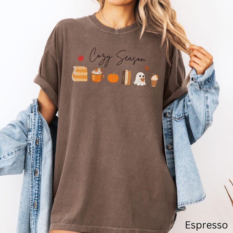 May include: Espresso-colored t-shirt with the text "Cozy Season" and fall-themed graphics. The design includes stacks of books, a pumpkin spice latte, a pumpkin, a book, a ghost, and a fall-themed drink.