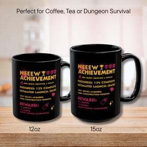 May include: Two black ceramic mugs with the text "NEEEW ACHIEVEMENT" in pink and yellow. The mugs have a capacity of 12oz and 15oz. The text includes details about a side quest, progress, and rewards.