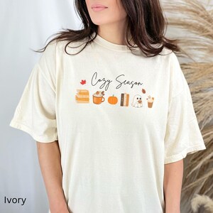 May include: Ivory-colored t-shirt with the text "Cozy Season" and fall-themed graphics. The design includes a stack of books, a pumpkin spice latte, a pumpkin, a book, a ghost, and a coffee cup.