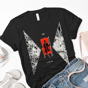 Op de afbeelding: Zwart T-shirt met een witte en zwarte afbeelding van een vrouw die op een rode troon zit met een kroon en een kaartontwerp. De tekst "A Novel" is onder de afbeelding gedrukt.