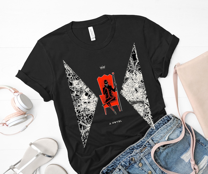Op de afbeelding: Zwart T-shirt met een witte en zwarte afbeelding van een vrouw die op een rode troon zit met een kroon en een kaartontwerp. De tekst "A Novel" is onder de afbeelding gedrukt.
