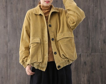 May include: A mustard yellow corduroy jacket with a button closure and large pockets. The jacket has a relaxed fit and is worn over a striped shirt and black trousers.
