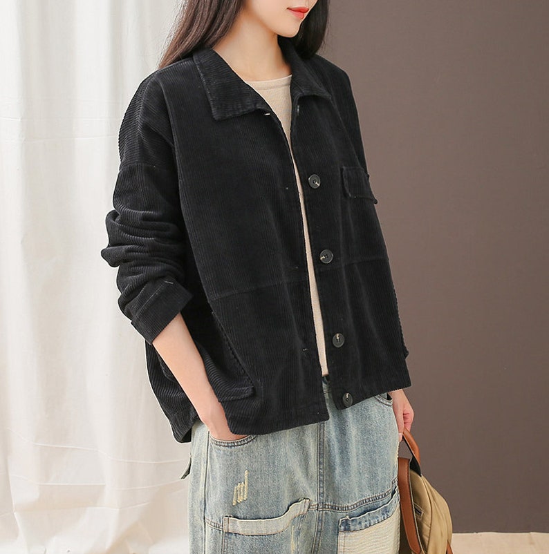 May include: A black corduroy jacket with a button-up front and a patch pocket on the right side. The jacket is worn over a white shirt and blue jeans.