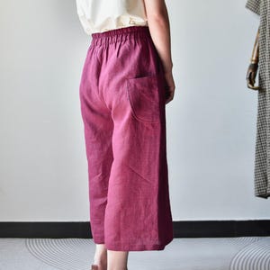 Puede incluir: Una persona lleva una camiseta sin mangas blanca y pantalones de lino de color magenta de pierna ancha con un solo bolsillo. Los pantalones tienen una cintura el&aacute;stica. Se usan zapatos de cuero marr&oacute;n. Un maniqu&iacute; con un vestido a cuadros est&aacute; en el fondo.