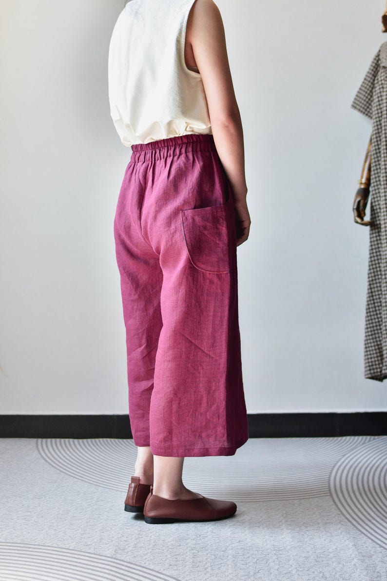 Puede incluir: Una persona lleva una camiseta sin mangas blanca y pantalones de lino de color magenta de pierna ancha con un solo bolsillo. Los pantalones tienen una cintura el&aacute;stica. Se usan zapatos de cuero marr&oacute;n. Un maniqu&iacute; con un vestido a cuadros est&aacute; en el fondo.
