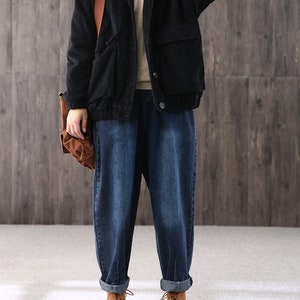May include: A black corduroy jacket with a brown leather strap and a large pocket on the front. The jacket is worn over a beige sweater and blue jeans.