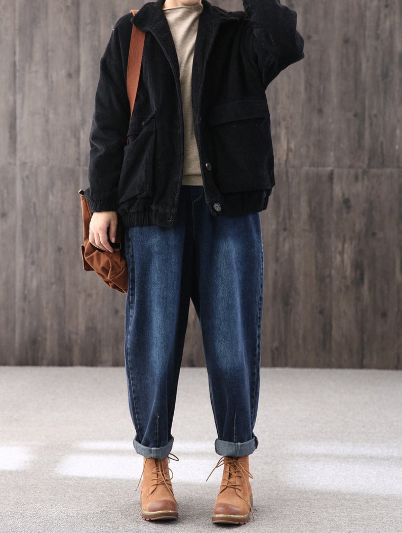 May include: A black corduroy jacket with a brown leather strap and a large pocket on the front. The jacket is worn over a beige sweater and blue jeans.