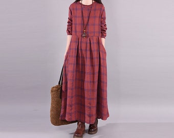 Puede incluir: Un vestido largo a cuadros rojos con mangas largas y bolsillos. El vestido tiene un cuello redondo y se complementa con un collar con colgante. Un bolso tote tejido y botas marrones completan el conjunto.