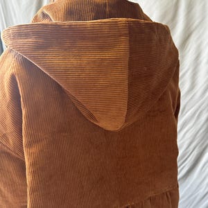 May include: A brown corduroy hooded jacket with a long, loose fit. The jacket has a drawstring hood and a relaxed, casual style.