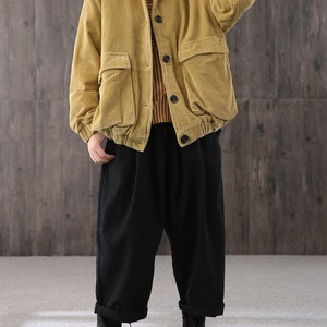 May include: A mustard yellow corduroy jacket with a button closure and large pockets. The jacket has a relaxed fit and is worn over a striped shirt and black pants.