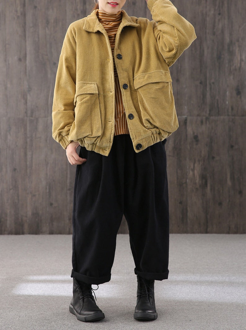 May include: A mustard yellow corduroy jacket with a button closure and large pockets. The jacket has a relaxed fit and is worn over a striped shirt and black pants.