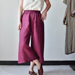 Puede incluir: Una camiseta sin mangas de color crema combinada con pantalones de pierna ancha burdeos. Los pantalones tienen una cintura el&aacute;stica. Zapatos de cuero marr&oacute;n completan el conjunto. Un vestido con un estampado de cuadros marrones y blancos es visible en el fondo.