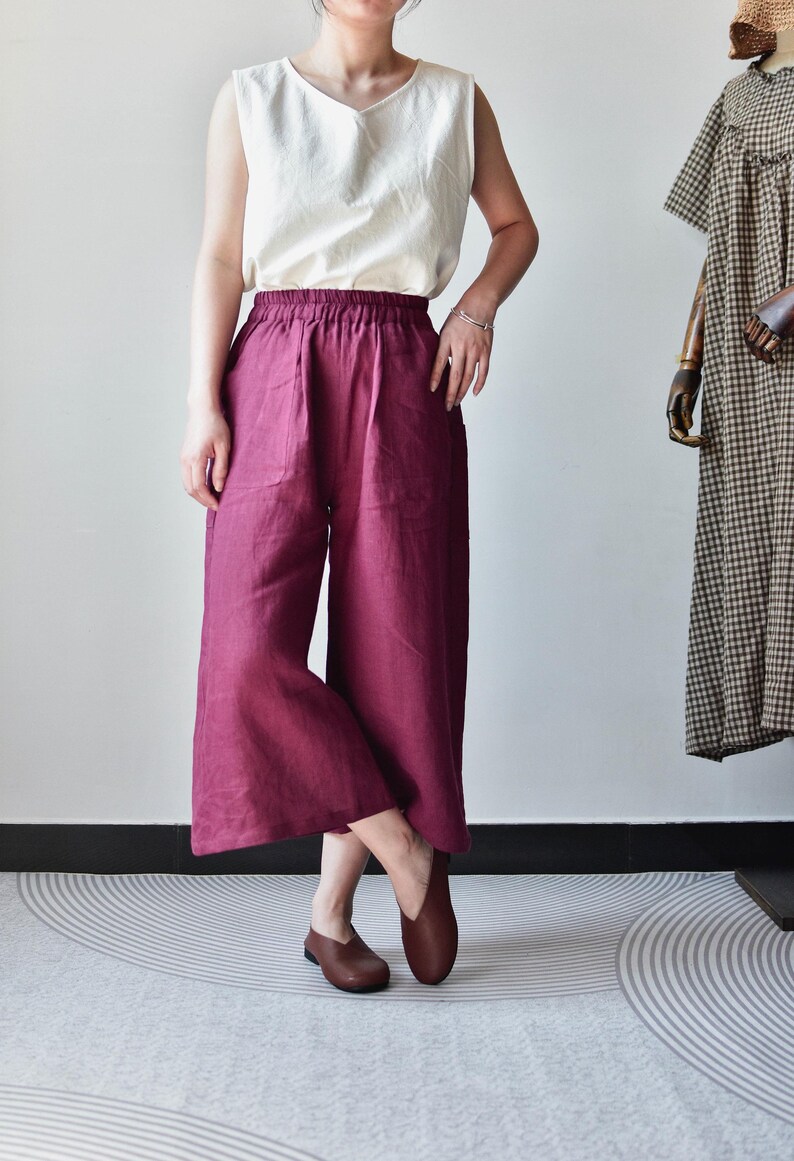 Puede incluir: Una camiseta sin mangas de color crema combinada con pantalones de pierna ancha burdeos. Los pantalones tienen una cintura el&aacute;stica. Zapatos de cuero marr&oacute;n completan el conjunto. Un vestido con un estampado de cuadros marrones y blancos es visible en el fondo.