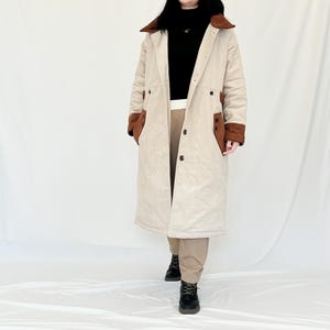 Puede incluir: Un abrigo largo de pana beige con detalles en ante marrón en el cuello y las mangas. El abrigo tiene cierre de botones y un corte holgado.