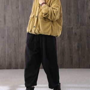 May include: A mustard yellow corduroy jacket with a button-up front and a patch pocket on the left side. The jacket is worn over a black turtleneck and black pants.