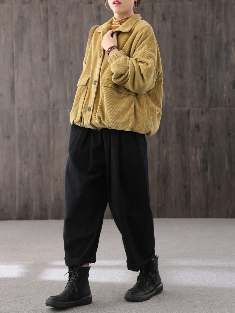 May include: A mustard yellow corduroy jacket with a button-up front and a patch pocket on the left side. The jacket is worn over a black turtleneck and black pants.