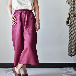 Puede incluir: Una persona viste un top sin mangas color crema y pantalones anchos burdeos. Los pantalones tienen cintura alta y bolsillos. Zapatos de cuero marr&oacute;n completan el conjunto. Un vestido en un maniqu&iacute; est&aacute; en el fondo.