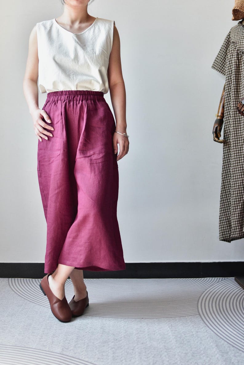 Puede incluir: Una persona viste un top sin mangas color crema y pantalones anchos burdeos. Los pantalones tienen cintura alta y bolsillos. Zapatos de cuero marr&oacute;n completan el conjunto. Un vestido en un maniqu&iacute; est&aacute; en el fondo.