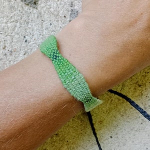 Op de afbeelding: Een geweven groene armband met zwarte touwtjes. De armband is gemaakt van een textuurrijk materiaal. De armband wordt om een pols gedragen.