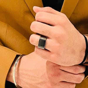 Puede incluir: Un anillo plateado con una piedra negra rectangular se lleva en una mano. Una pulsera dorada está en la muñeca de la misma mano. Una blazer marrón se lleva en el fondo. Un reloj con una correa de cuero marrón está en la otra muñeca.