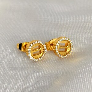 Op de afbeelding: Een paar goudkleurige oorstekers. Elke oorbel heeft een cirkelvormig ontwerp met een centrale, open vorm en een omringende rand van kleine, heldere edelstenen. De oorbellen zijn geplaatst tegen een witte, getextureerde achtergrond.