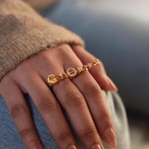 Könnte beinhalten: Goldfarbene Ringe mit dem Wort "LOVE", mit einem diamantartigen Detail auf dem "O". Die Ringe werden an einer Hand mit polierten Nägeln getragen. Die Ringe haben ein zartes, elegantes Design.