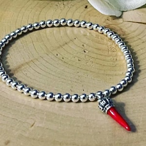 Puede incluir: Un brazalete de plata con cuentas y un dije de cuerno rojo. El brazalete está hecho de cuentas pequeñas y redondas y tiene un diseño simple y elegante.