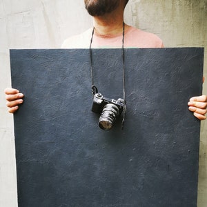 May include: A person holding a large, dark grey rectangular board in front of their body. A black camera with a lens is hanging from a strap around their neck.