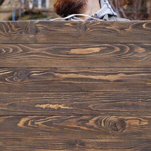 May include: A rectangular wooden sign with a dark brown finish. The sign is made of horizontal planks with visible wood grain and knots. The sign is being held by a person.