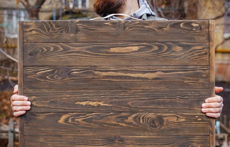 May include: A rectangular wooden sign with a dark brown finish. The sign is made of horizontal planks with visible wood grain and knots. The sign is being held by a person.