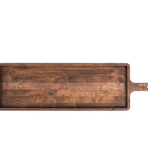 Op de afbeelding: Een lange, rechthoekige houten serveerschaal met een handvat aan &eacute;&eacute;n kant. De schaal is gemaakt van donkerbruin hout met een natuurlijke houtnerf.