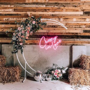 May include: A white metal moon archway decorated with pink and white flowers. The archway is set against a rustic wooden wall with a pink neon sign that reads "Crazy in Love".