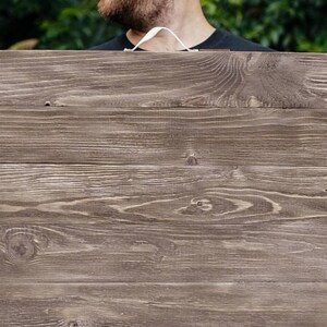 May include: A rectangular wooden sign with a dark brown finish. The sign is made of horizontal wooden planks and has a visible wood grain texture. A white cord is attached to the top. The sign is held by a person.