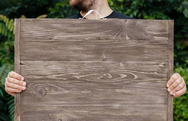 May include: A rectangular wooden sign with a dark brown finish. The sign is made of horizontal wooden planks and has a visible wood grain texture. A white cord is attached to the top. The sign is held by a person.