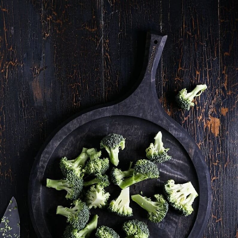May include: A black wooden serving board with a handle, featuring the text "WOODSTUFF" on the handle. The board is filled with fresh, green broccoli florets.