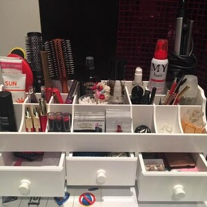 K&ouml;nnte beinhalten: Wei&szlig;er Organizer mit mehreren F&auml;chern zur Aufbewahrung von Make-up und Beauty-Produkten. Der Organizer hat drei Schubladen und ist mit verschiedenen Artikeln gef&uuml;llt, darunter B&uuml;rsten, Flaschen und Tuben.