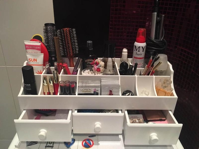 K&ouml;nnte beinhalten: Wei&szlig;er Organizer mit mehreren F&auml;chern zur Aufbewahrung von Make-up und Beauty-Produkten. Der Organizer hat drei Schubladen und ist mit verschiedenen Artikeln gef&uuml;llt, darunter B&uuml;rsten, Flaschen und Tuben.
