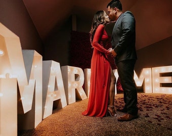Puede incluir: Una pareja se abraza y se besa frente a grandes letras blancas iluminadas que deletrean "Marry Me". La mujer lleva un vestido rojo y el hombre lleva un traje oscuro. El fondo está decorado con pétalos de rosas rojas.
