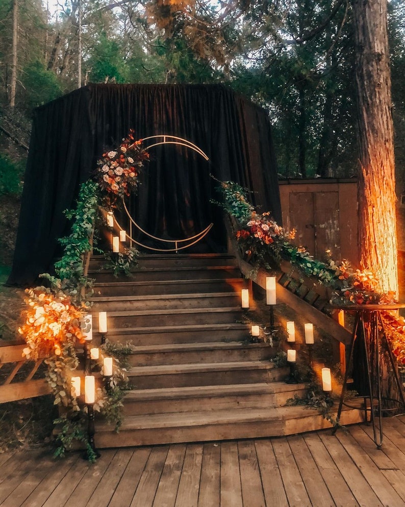 May include: A wooden staircase with a black fabric backdrop and a gold crescent moon archway. The staircase is decorated with greenery and candles.