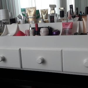 K&ouml;nnte beinhalten: Wei&szlig;er Holzorganizer mit drei Schubladen und einem oberen Regal mit F&auml;chern zur Aufbewahrung von Kosmetik und Toilettenartikeln. Der Organizer hat ein schlichtes Design mit runden Kn&ouml;pfen an den Schubladen.