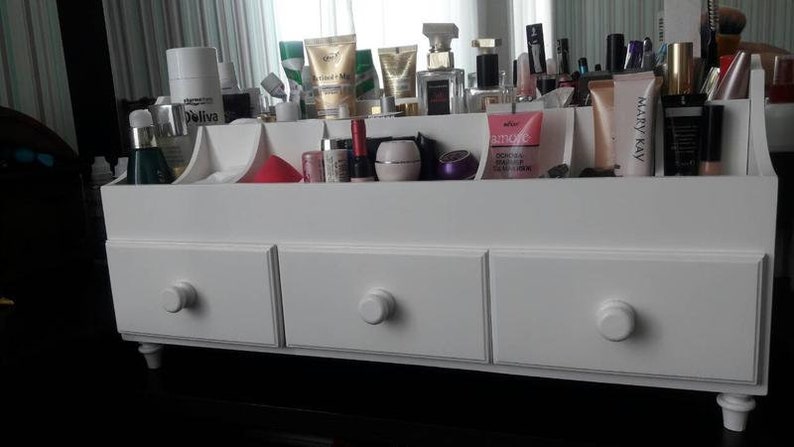 K&ouml;nnte beinhalten: Wei&szlig;er Holzorganizer mit drei Schubladen und einem oberen Regal mit F&auml;chern zur Aufbewahrung von Kosmetik und Toilettenartikeln. Der Organizer hat ein schlichtes Design mit runden Kn&ouml;pfen an den Schubladen.