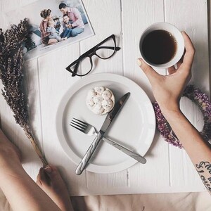 May include: A white plate with a fork and knife, a small white cake with a brown topping, and a cup of coffee on a white wooden surface. A person's arm with a tattoo is visible in the background.